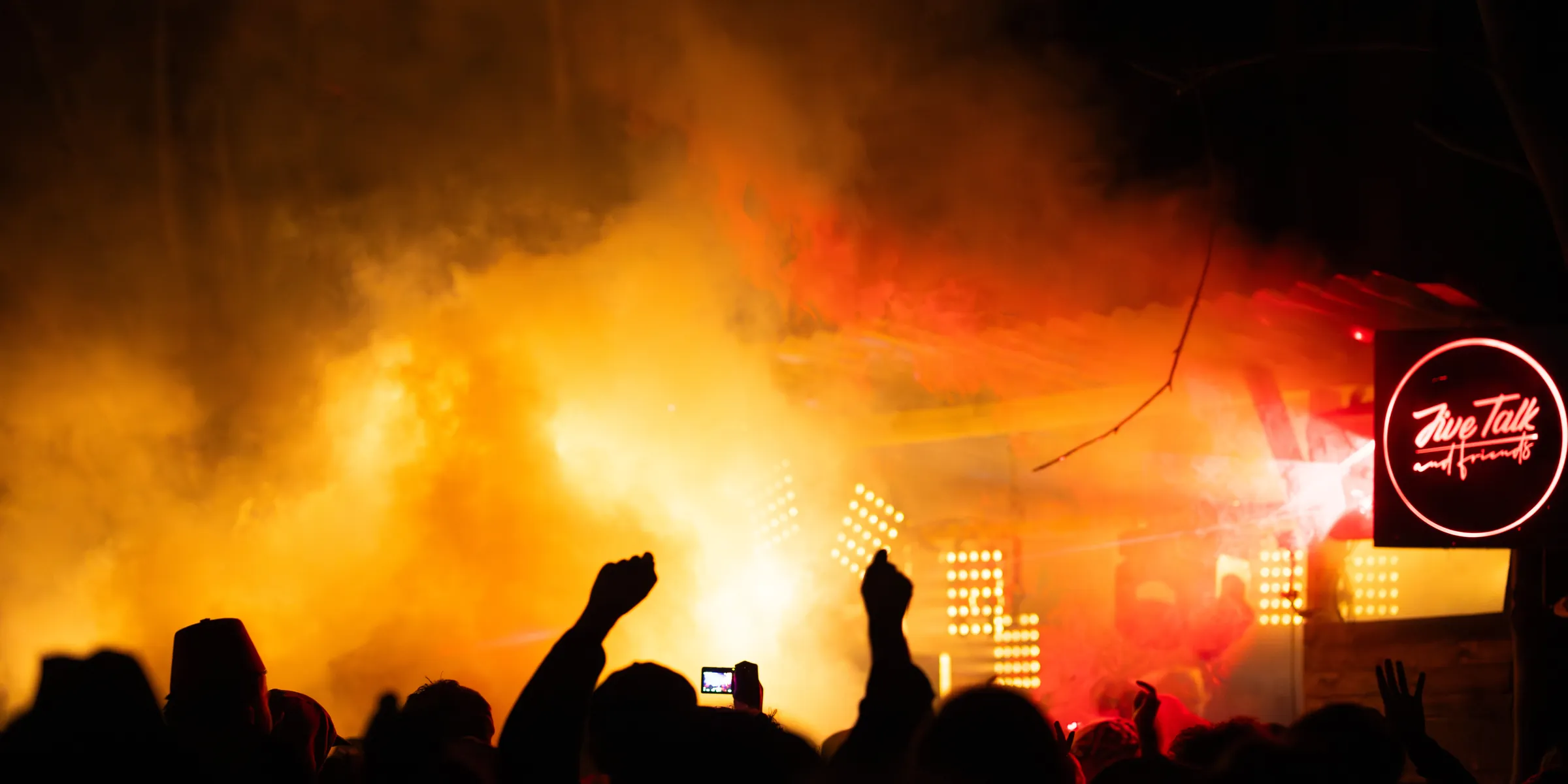 A festival crowd in silhouette, fists raised towards a stage lit amber and red through thick smoke